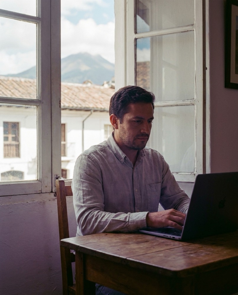 Estudiante rindiendo un examen en línea en su laptop, en Quito, con casas coloniales y el volcán Pichincha por la ventana