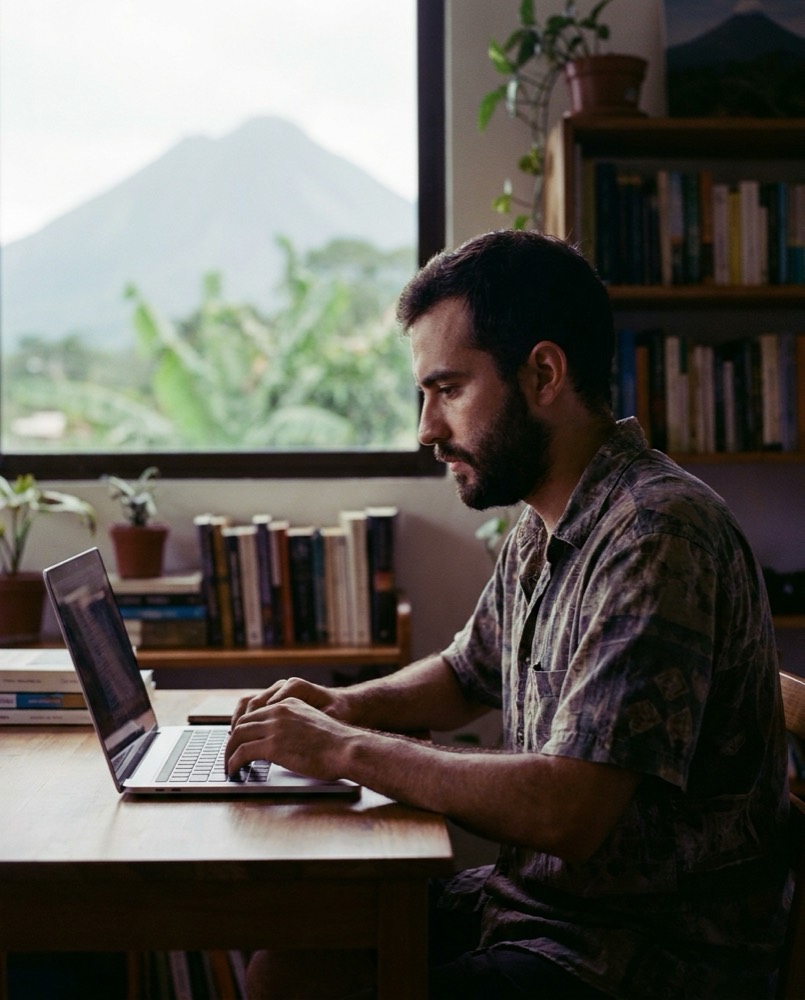 Estudiante rindiendo un examen en línea en su laptop, en San Salvador, con el volcán de San Salvador por la ventana