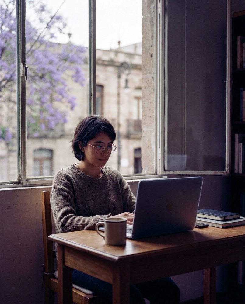 Estudiante rindiendo un examen en línea en su laptop, en Ciudad de México, con jacarandas en flor por la ventana